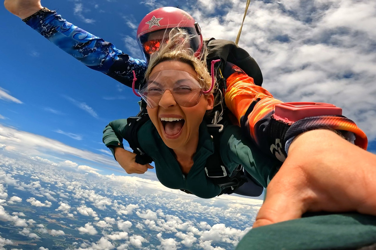 A woman with goggles smiles widely while tandem skydiving, with her instructor behind her. They are free-falling above clouds under a blue sky, with the ground far below.